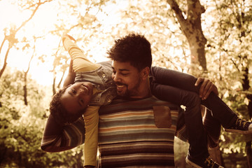 Canvas Print - Smiling African American father in park carrying his daughter on shoulders.