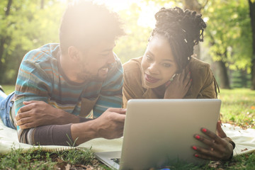 Sticker - Cheerful African American couple lying down in park and using laptop.