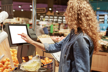 Back view of curly unrecognizable curly woman in denim jacket, weighs fruit products on electronic scales with touch screen, poses in supermarket, buys bananas. Now shopping in easier for you