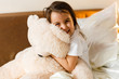 © Angelov - Sweet little girl is hugging a teddy bear looking at camera and smiling while sitting on her bed 396815581