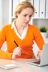 Young beautiful business woman or student communicates by laptop computer while sitting at the desk and holding black pen. She Orange sweater is at the girl. Education, business and lifestyle concept