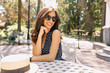 © PhotoBook - Image of young woman with beautiful hair and charming smile is sitting in summer cafeteria in sunlight. She is wearing pretty summer dress and black sunglasses. Background park. Beautiful portrait.