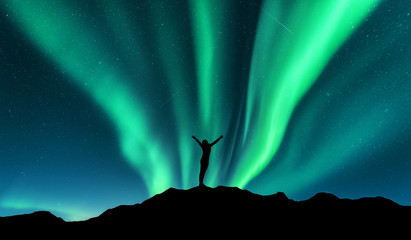 Wall Mural - Aurora and silhouette of standing woman with raised up arms on the mountain in Norway. Aurora borealis and happy girl. Starry sky, green polar lights. Night landscape. Northern lights. Travel