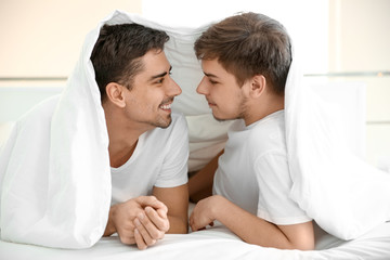 Poster - Young gay couple lying under blanket on bed in light room