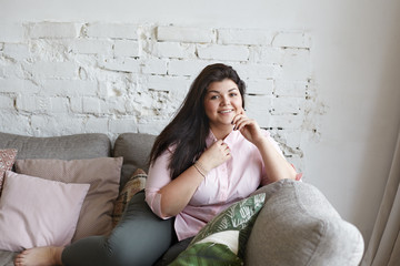 picture of body positive charming young woman posing indoors on gray couch against white brick wall 
