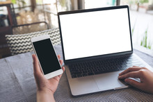 Mockup Image Of Hands Holding Blank Mobile Phone While Using Laptop With Blank White Desktop Screen On Table