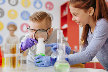 Wall Mural - Examining thoroughly. Joyful teenage boy examining a chemical substance in the flask with a magnifying glass while his classmate holding that flask for him