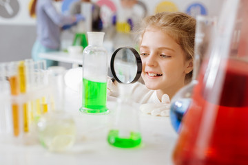 Wall Mural - Helpful device. Cute pretty teenage girl squatting near the table in the chemical laboratory and checking for sediment in the flask with the help of a magnifying glass