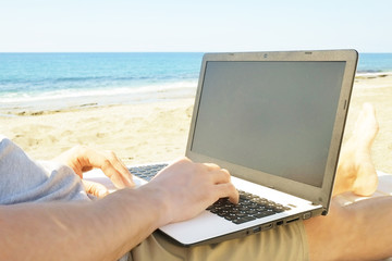 Fit young man sitting on deck chair at sea view beach using laptop, blank screen. Male freelance programmer in chaise-long lounge working coding blogging surfing on notebook computer. Distance work co