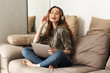 © Drobot Dean - Image of pleased woman 20s with brown hair listening to music using wireless headphones and laptop, while sitting on sofa in living room with legs crossed