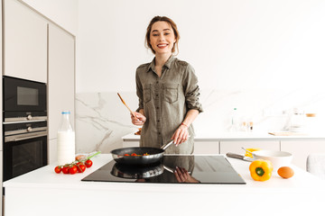 Portrait of positive smiling woman cooking healthy breakfast in kitchen, frying ingredients on pan