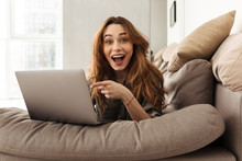 Image Of Happy European Woman Lying On Sofa In Living Room, And Pointing Finger On Screen Of Notebook With Interest Or Surprise