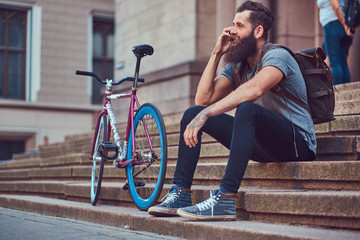 A handsome hipster traveler with a stylish beard and tattoo on his arms dressed in casual clothes, sitting on the steps, using the phone, resting after a bike ride.