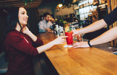 Attractive and gorgeous woman sitting at the stand and getting her order from barman. It is red cocktail. She looks to barman and smiling. Also there is another customer sitting not very far from her.