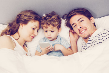 Wall Mural - Portrait of happy young Caucasian parents lying in bed with their little son. Little boy using mobile phone