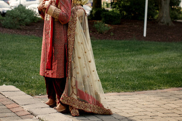 Indian fiance in red wedding costume holds bride's hands on his chest while she hugs him from behind