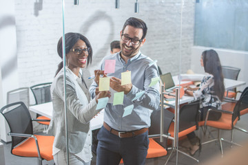 Smiling woman and man discussing ideas and brainstorming with sticky notes on glass wall in office