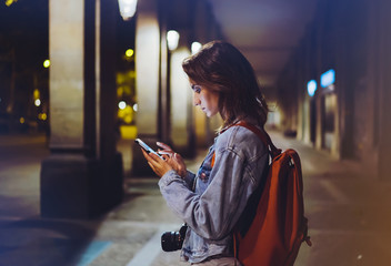 Wall Mural - blogger hipster using in hands gadget mobile phone, woman with backpack pointing finger on blank screen smartphone on background bokeh light in night atmospheric city, online wifi internet concept