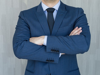 Torso of a businessman standing with folded arms in a classic navy blue suit against grey backgound.