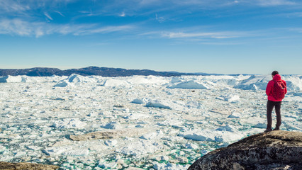 Wall Mural - Travel in arctic landscape nature with icebergs - Greenland tourist man explorer - tourist person looking at amazing view of Greenland icefjord - aerial video. Man by ice and iceberg in Ilulissat.