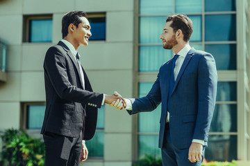 Wall Mural - Closeup of two smiling business men shaking hands with office building in background. Side view.