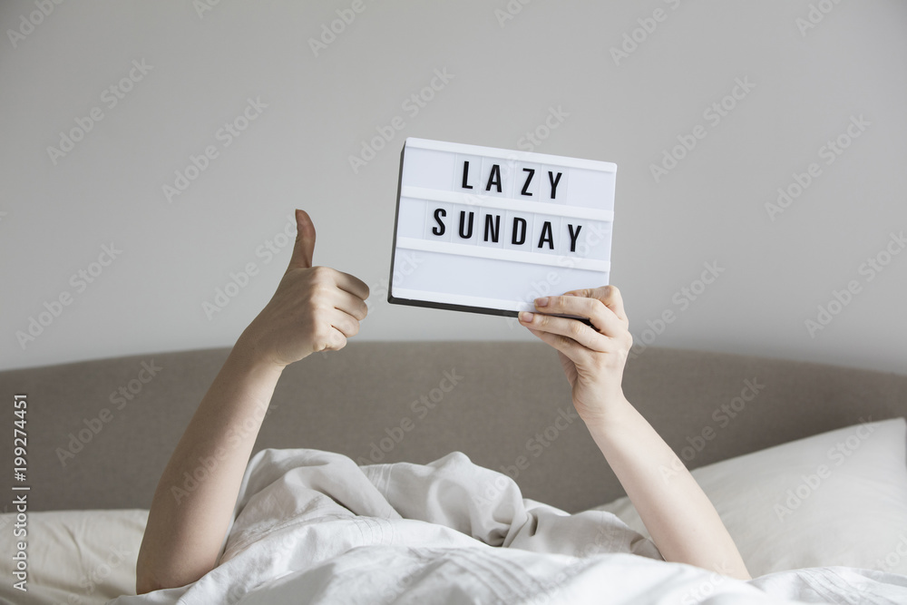 Female in bed under the sheets holding up a lazy sunday sign - obrazy, fototapety, plakaty 