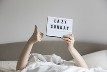 Female In Bed Under The Sheets Holding Up A Lazy Sunday Sign