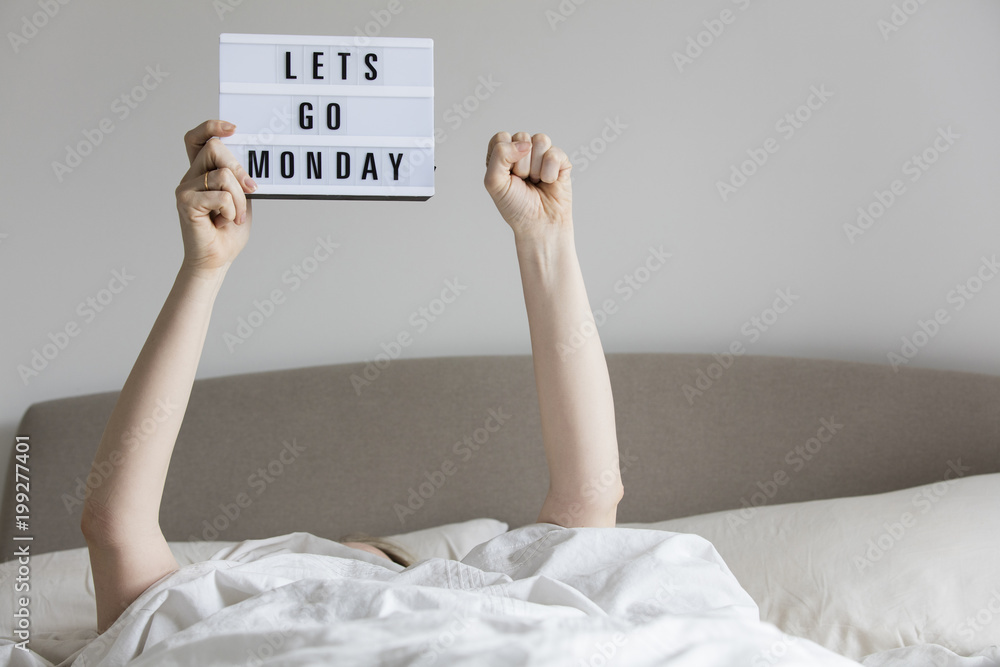 Female in bed under the sheets holding up a lets go monday sign - obrazy, fototapety, plakaty 