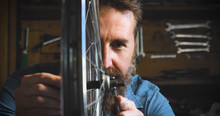 Well-groomed Handsome Bearded Master Hipster, Specialist In Bicycles, Repairing A Bicycle In His Workshop, Wheels, Frame, Spokes, The Background Of Tools. Concept: Pro Bike, Cycle Passion, Lifestyle.