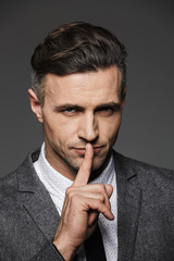 Sticker - Photo of trendy man dressed in businesslike suit looking on camera and showing silence gesture with finger on lips, isolated over gray background