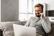 © Drobot Dean - Image of positive man 30s in casual wear sitting on sofa in living room, while using laptop and speaking on cell phone