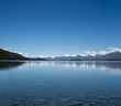 © josemanuelerre - Imagen de un paisaje azul minimalista con montañas nevadas distantes y un lago enorme y claro. Las montañas se reflejan en el agua. Y hay muchas rocas y piedras en el fondo del lago.