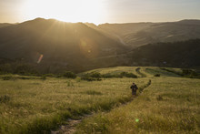 High Angle View Of Man On Trail Amidst Grassy Field Against Mountains During Sunset