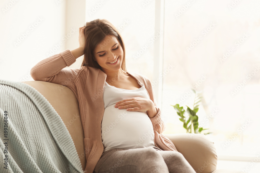 Young pregnant woman sitting on couch in living room - obrazy, fototapety, plakaty 