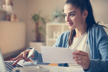 Smiley young girl at home holding document and typing on laptop. Close up.