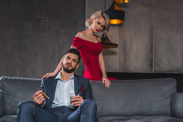 man in suit holding cigar and glass of whisky while spending time with sexy girl in red dress