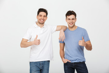 Wall Mural - Two happy buddies 30s wearing casual t-shirt and jeans smiling and showing thumbs up on camera in studio, isolated over white background