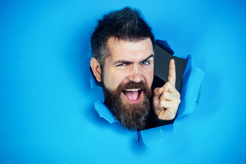 Wall Mural - Bearded man making hole in paper. View of male face through hole in blue paper. Bearded man looking through hole and show the finger. Bearded man making hole in paper and points up.