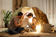 Hygge And People Concept - Father With Torch Light Telling Scary Stories To His Daughter And Wife, Family Having Fun In Kids Tent At Night At Home