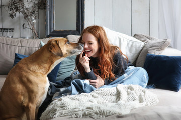 Hübsche rothaarige Frau sitzt auf einem Sofa lacht und wird von einem Hund angestupst