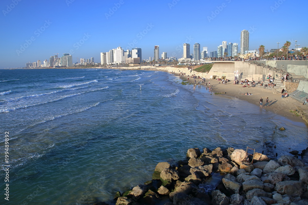 Zatoka Morza Śródziemnego w Tel Awiwie, niebieska czysta woda, kamienista i piaszczysta plaża z turystami, na horyzoncie wyoskie, jane, nowoczesne budynki miasta, bezchmurne błękitne niebo - obrazy, fototapety, plakaty 