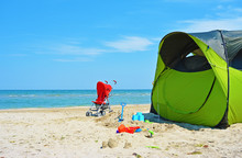 Summer Camping With A Tent At A Lonesome Wild Beach With A Turquoise Sea And Blue Sky In The Background, Baby Stroller And Toys. Camp Touristic With Kids.