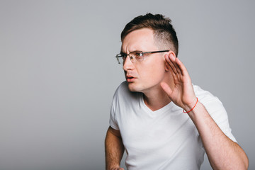 Curious handsome young man in  shirt overhearing rumors over grey background.