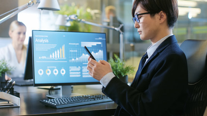 Wall Mural - In the Office Asian Businessman Uses Smartphone, Typing Important Message. His Desktop Computer Shows Sratistics with Company Growth. Stylish Office.