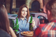 Friends having a drinks in a bar, They are sitting at a wooden table with beers and pizza. Focus on a gorgeous girl touching her bottle.