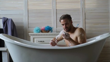 Wall Mural - Young handsome man relaxing in the tub. Man with beard having a bath in a white buthtub. Men covered with foam bubble bath in luxury bathtub with happiness and relaxing.