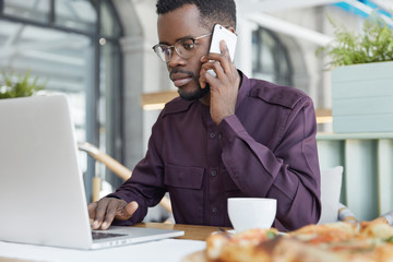 Wall Mural - Concentrated dark skinned man in formal wear looks confidently into laptop computer, has serious expression, solves financial problems during coffee break, reads important information in internet