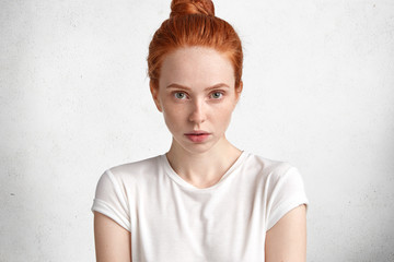 Confident adorable lovely young ginger female with attractive look, has pure skin, dressed in casual t shirt, poses against white concrete wall. Red haired woman student listens something attentively