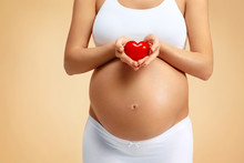 Close Up Of Pregnant Woman Holding Red Heart On Beige Background. Pregnancy, Maternity, Preparation And Expectation Concept
