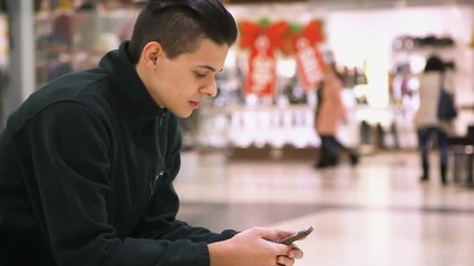 Wall Mural - Profile of stylish young man with crew haircut who sits and keeps a smartphone in his hands. He surfs the net in an airy shopping mall in autumn